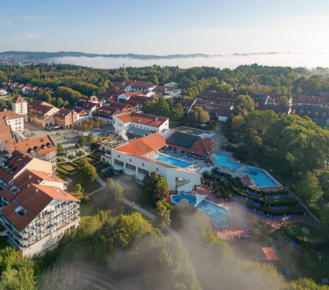 Luftaufnahme der Wohlfühltherme Bad Griesbach mit Außenbecken, umliegender Parklandschaft und Hotelgebäuden bei Morgennebel.